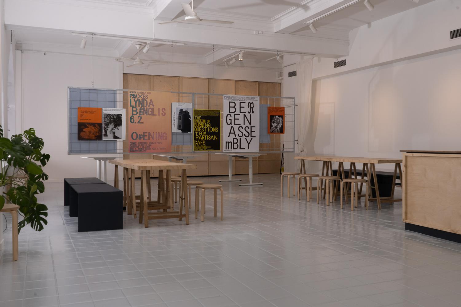 Minimalistisk gallerirom med hvite vegger, høyt tak og store buede vinduer. Trébord med tilhørende krakker står plassert i midten, sammen med noen svarte benker. Plakater med tydelig typografi og grafisk design er utstilt på nettingvegger langs bakveggen, og reklamerer for kunstarrangementer og utstillinger. En grønn potteplante står ved vinduet og tilfører et snev av farge til det rene, moderne interiøret.