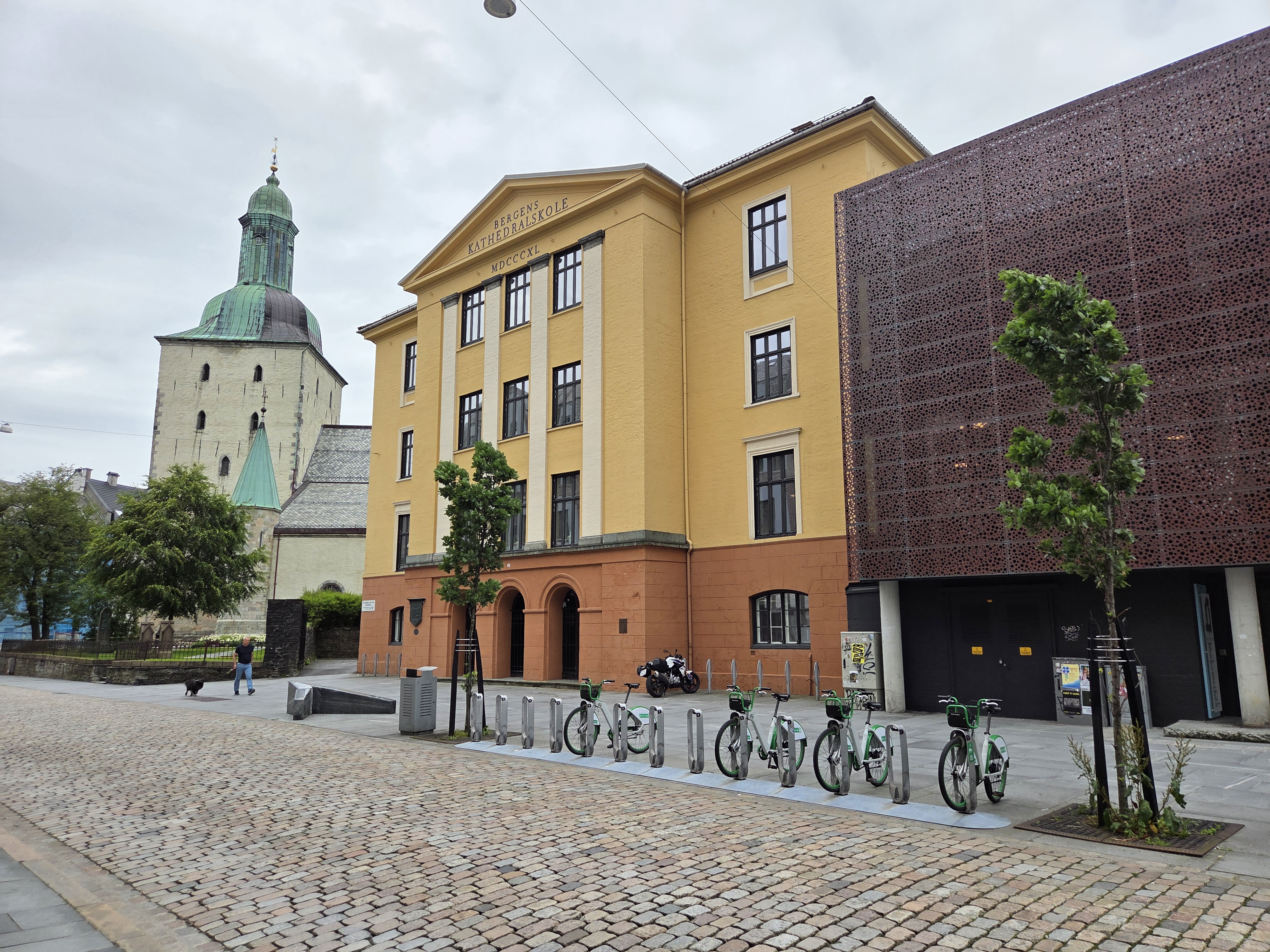 En historisk gul og oransje murbygning med klassiske detaljer, ved siden av det middelalderske steintårnet til Bergen Domkirke.