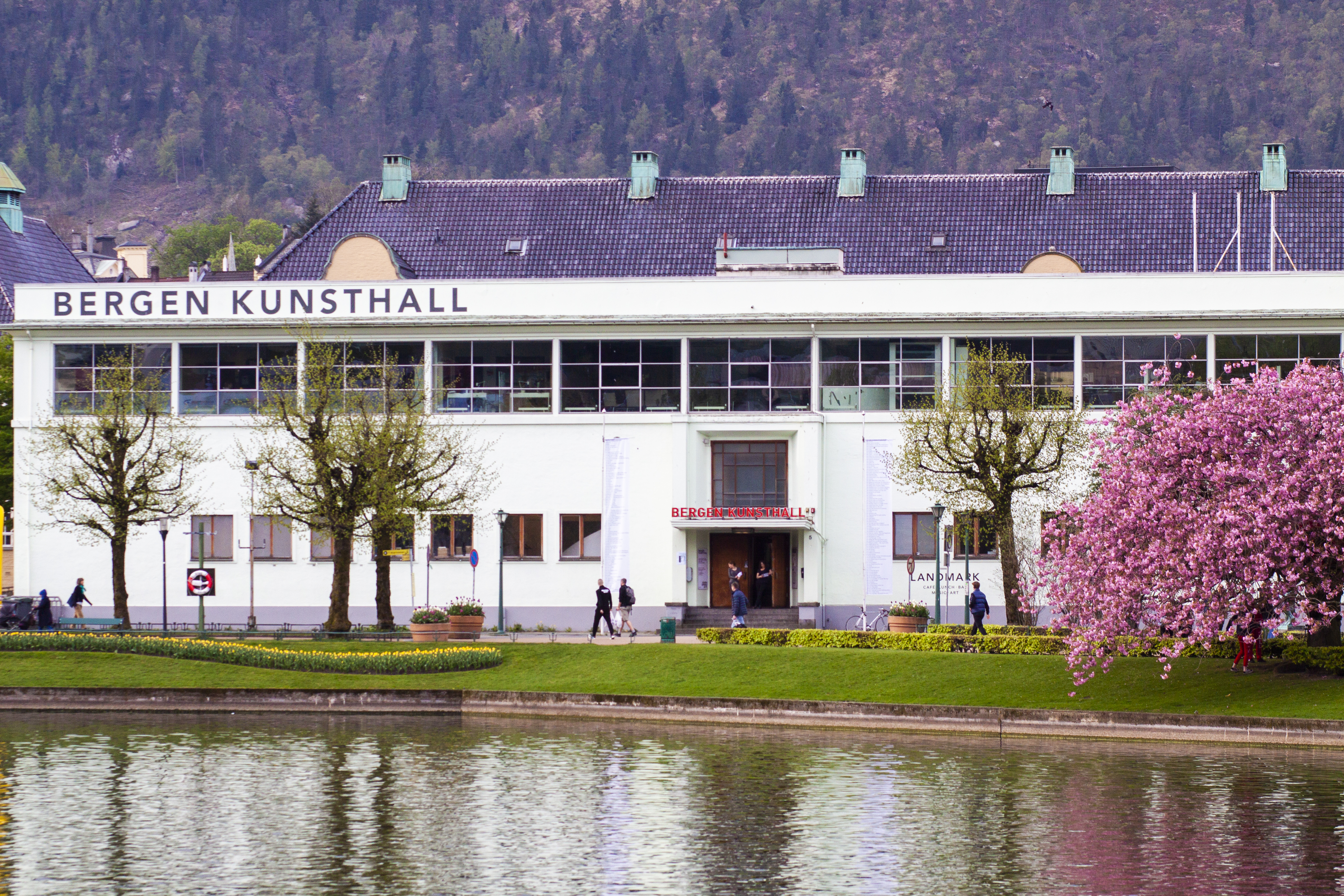 Fasaden til Bergen Kunsthall, en hvit modernistisk bygning med store horisontale vinduer og navnet «BERGEN KUNSTHALL» i tydelige bokstaver langs taklinjen. Inngangen er midtstilt med rød skrift, og bygget vender ut mot en park med trær og en dam i forgrunnen.
