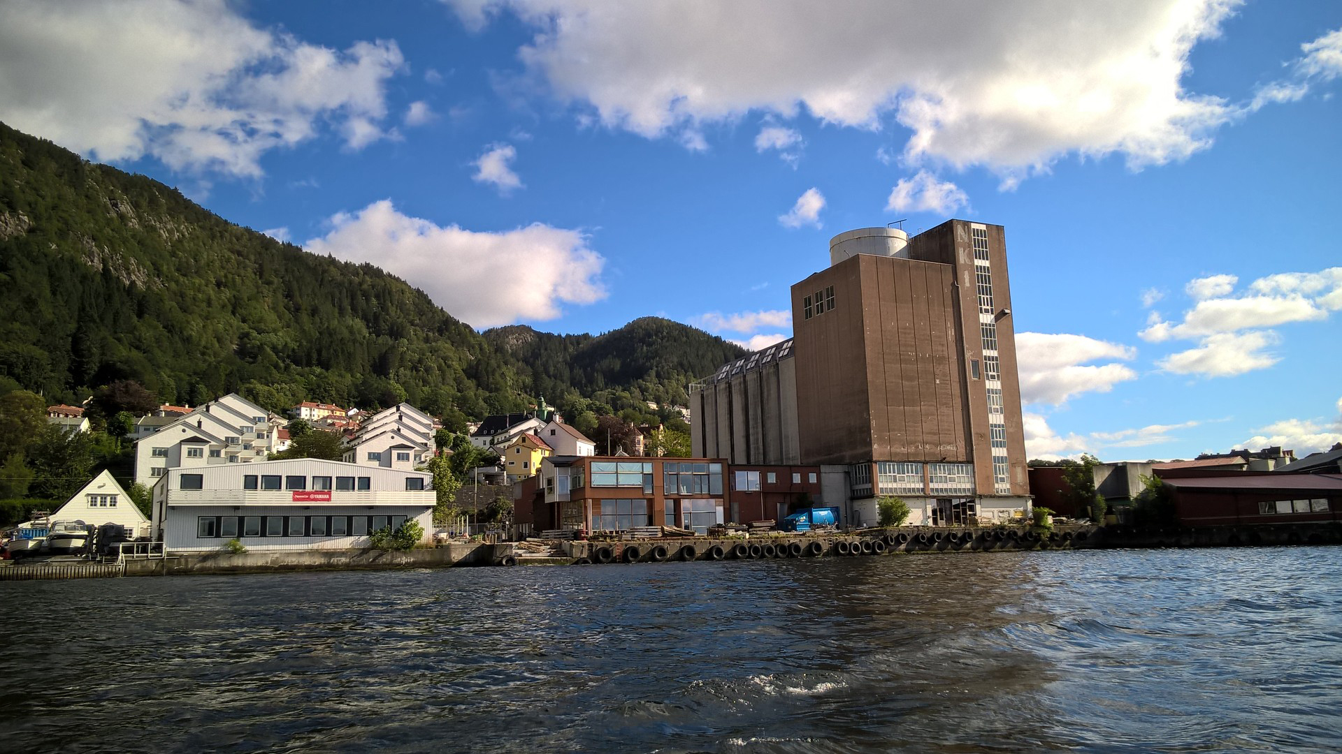 En stor, høy bygning i industriell stil med et tårn og tilhørende strukturer, plassert ved vannkanten med skogkledde åser i bakgrunnen.
