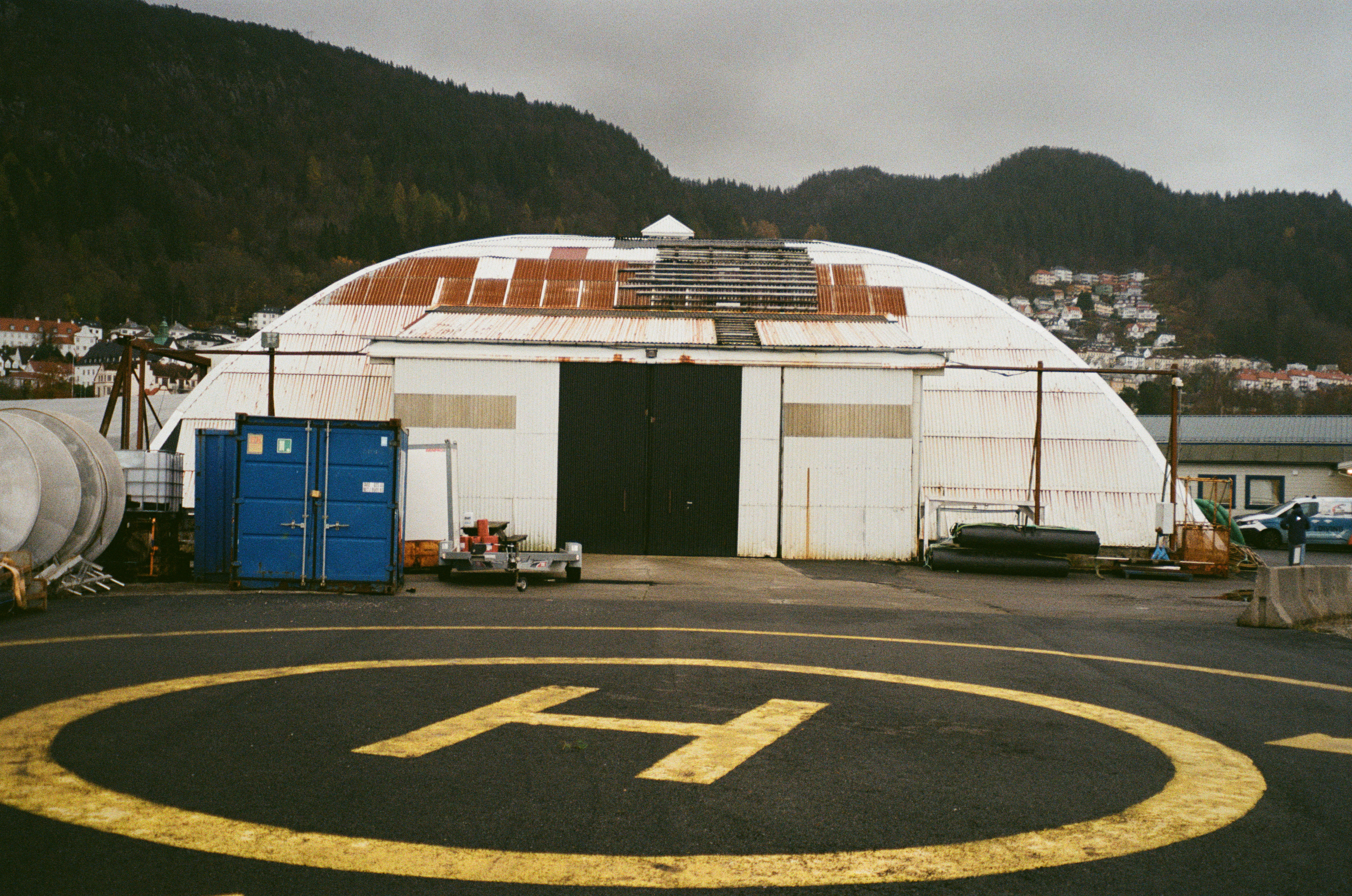 Gammelt hangar med rustent tak bak en helikopterplass merket med gult «H».