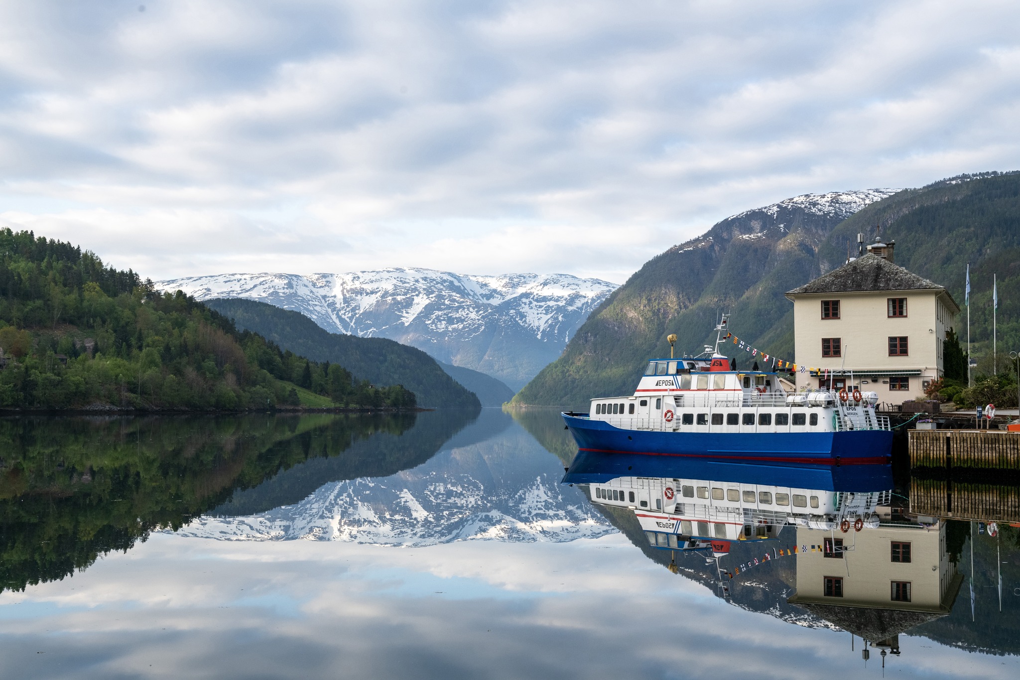 En hvit og blå passasjerbåt fortøyd ved en bygning ved en stille fjord.