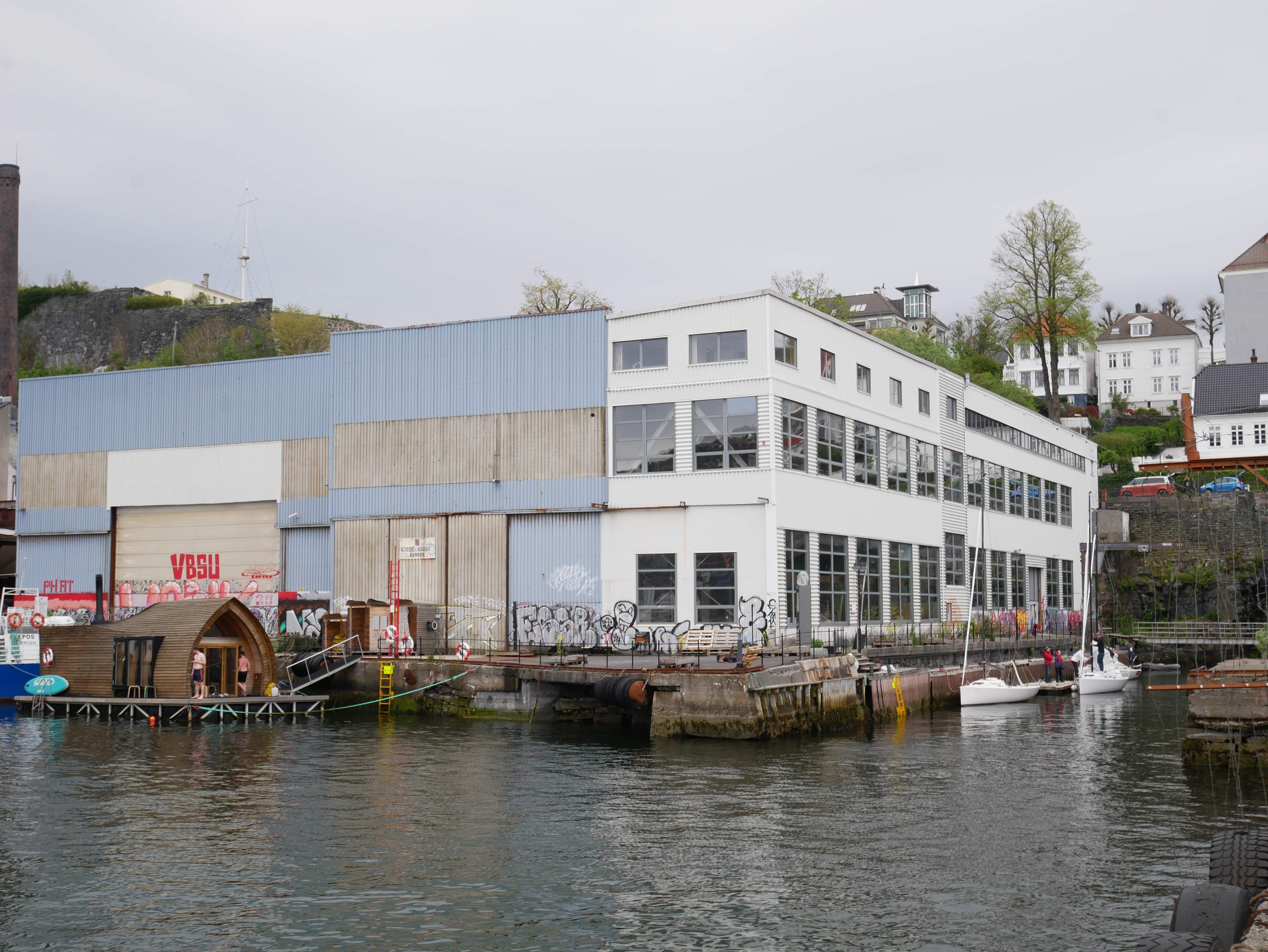 Stor industribygning ved vannkanten med graffiti på veggene og små båter fortøyd i nærheten.
